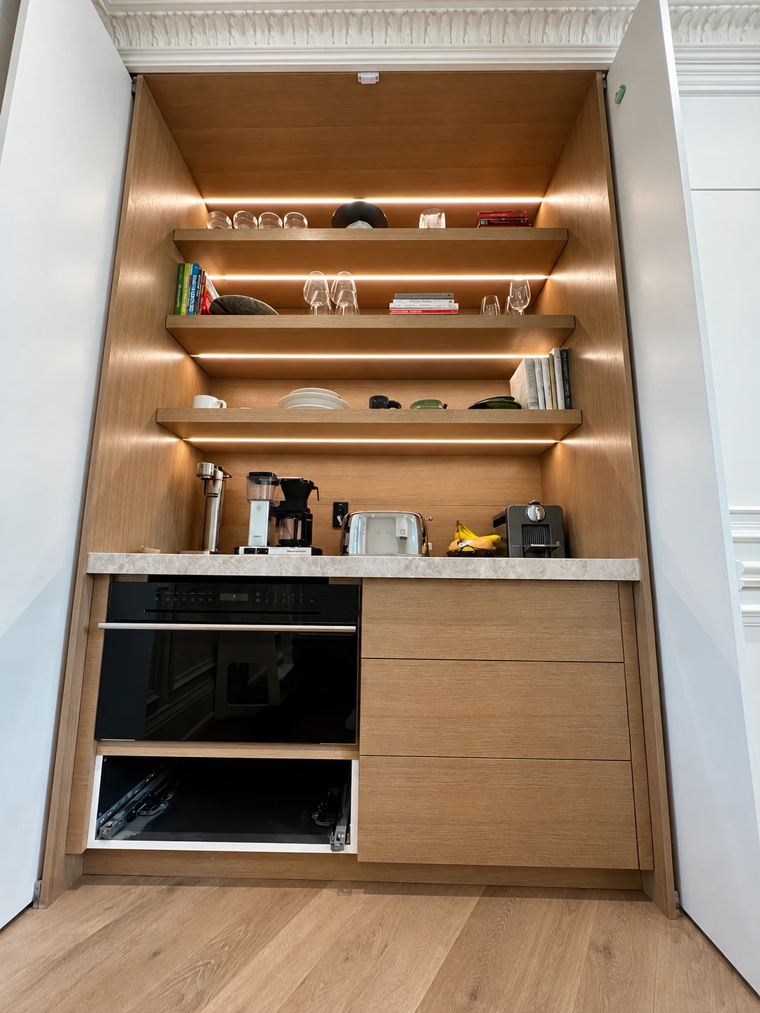 Concealed LED lighting illuminating wood shelves above a built-in oven and countertop appliances