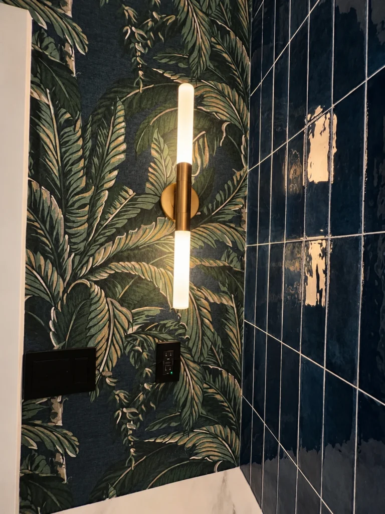 Brass double-ended tubular wall sconce beside botanical wallpaper and navy vertical tile