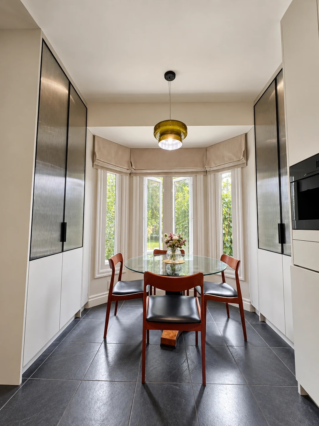 Amber glass pendant above a breakfast table between ribbed-glass cabinets and bay windows