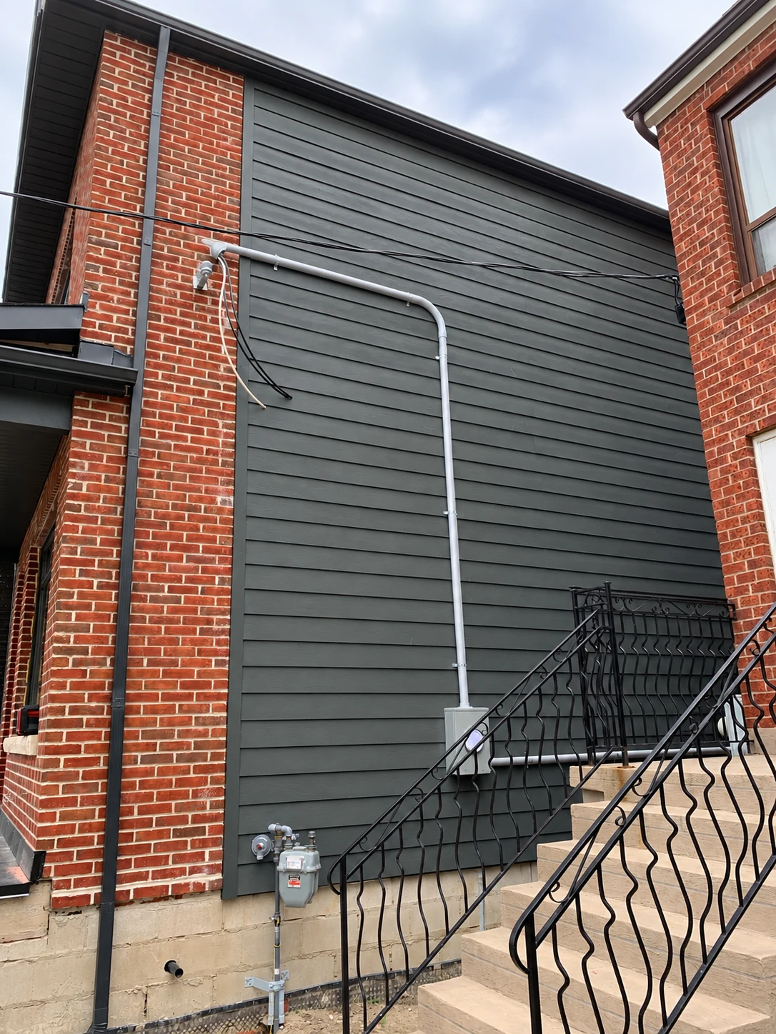 Exterior electrical service conduit routed along grey siding beside brick walls and stairs