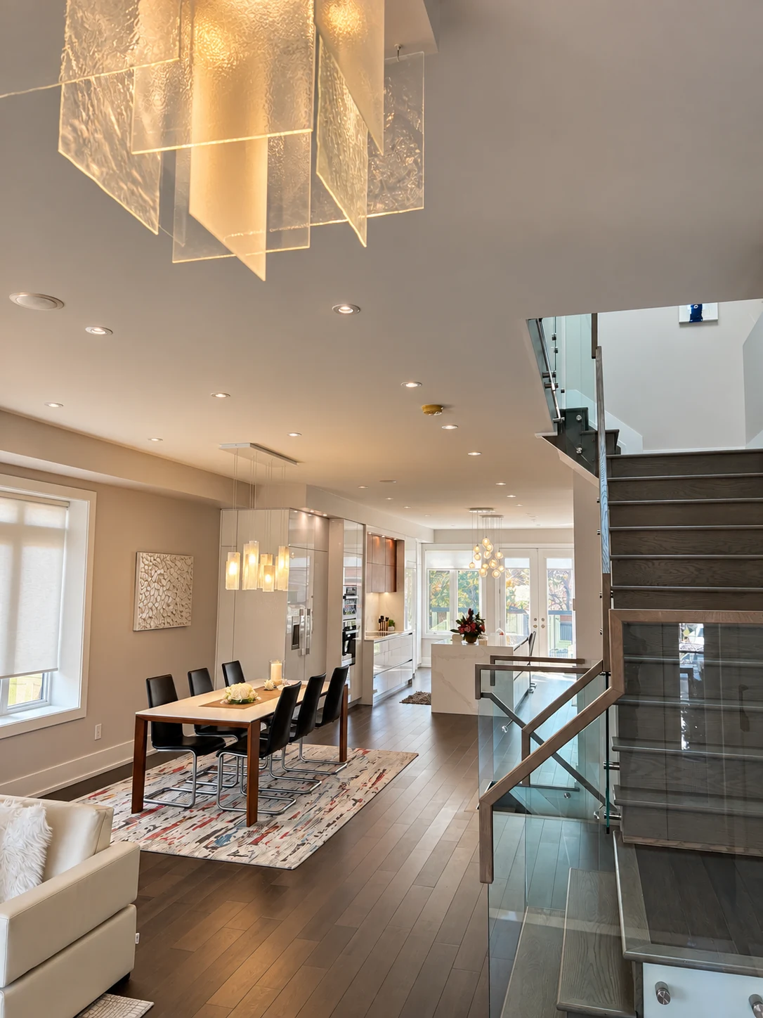 Chandeliers and recessed lighting across an open-plan home beside a glass staircase